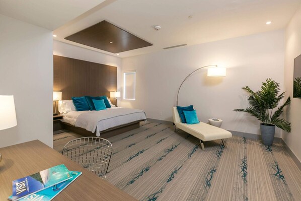 A modern hotel room with a bed, a desk, a chair, a sofa, a potted plant, and a wall-mounted lamp. - Suite 2, 15% Off Holiday Dates! (Cruz Bay)