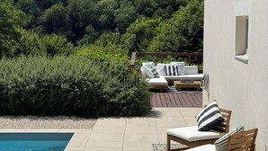 A poolside area with a view of a forested hillside, a wooden deck, and a white building with a window.