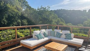 A wooden deck with a white sectional sofa and a wooden coffee table, surrounded by a wooden railing and lush greenery.