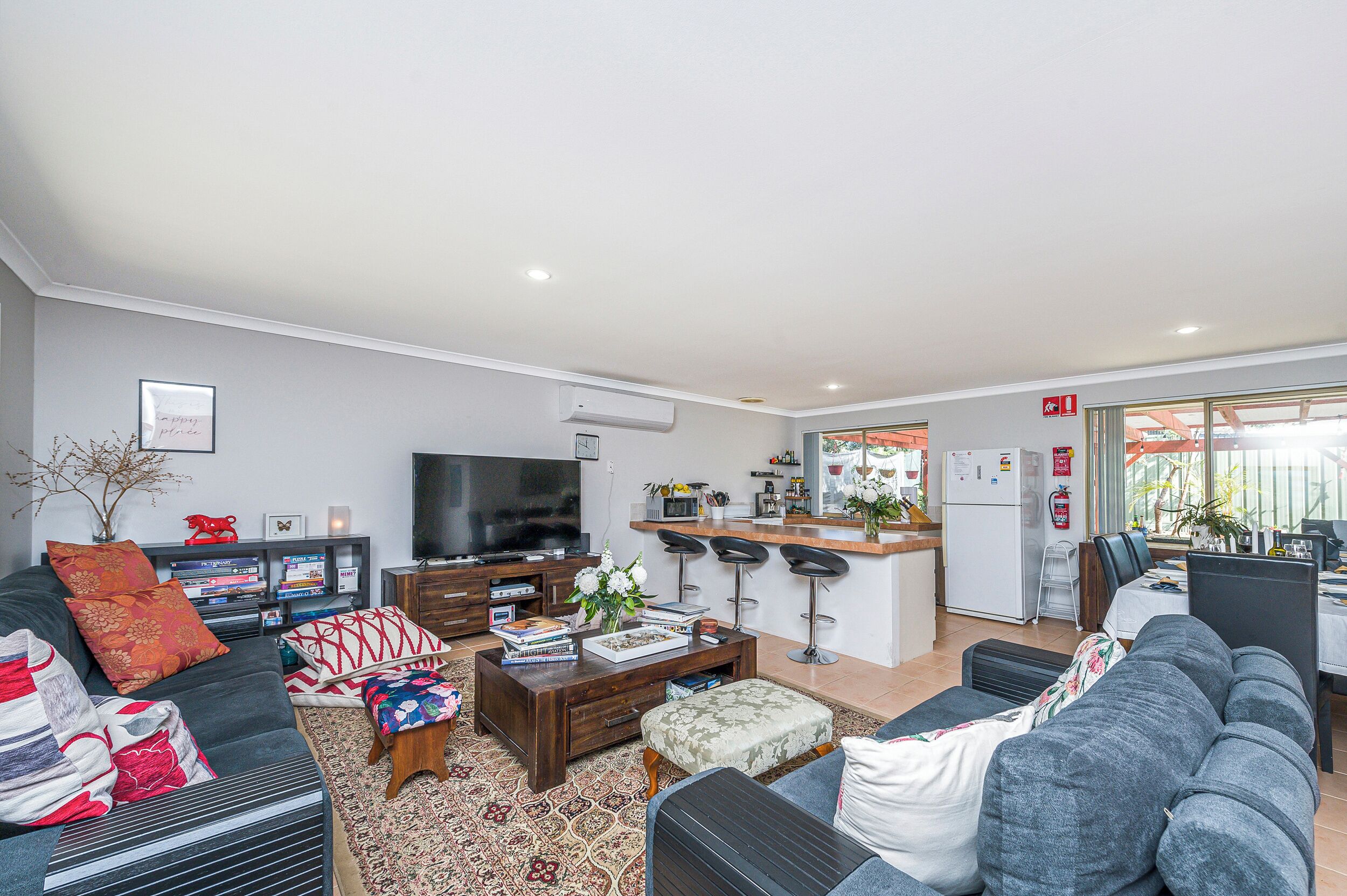 A modern living room with a sofa, coffee table, and TV. There's a kitchen area with a fridge and bar stools.