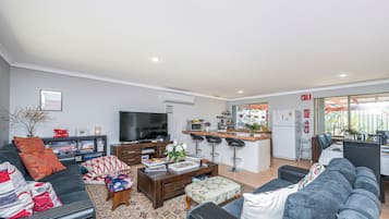 A modern living room with a sofa, coffee table, and TV. There's a kitchen area with a fridge and bar stools.
