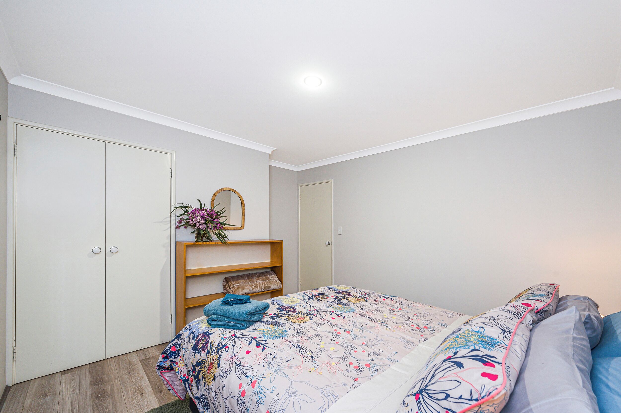 Une chambre à coucher avec un lit, une armoire, un miroir et une étagère sur laquelle est posé un vase de fleurs.