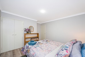 A bedroom with a bed, a wardrobe, a mirror, and a shelf with a vase of flowers. - Waves And Moore - Beautifully presented home (Guilderton)
