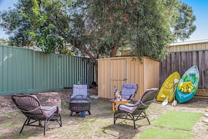 A backyard with a wooden shed, two wicker chairs, a small table with a bottle, and two kayaks.