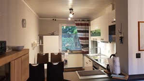 A compact kitchen with a dining area, a small refrigerator, a window with blinds, and a clock on the wall. - Comfortable vacation home in a central location in Bad Zwischenahn with garden (Bad Zwischenahn)