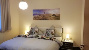 A neatly made bed with floral bedding, a wall-mounted painting of a coastal scene, and bedside lamps. - Comfortable vacation home in a central location in Bad Zwischenahn with garden (Bad Zwischenahn)