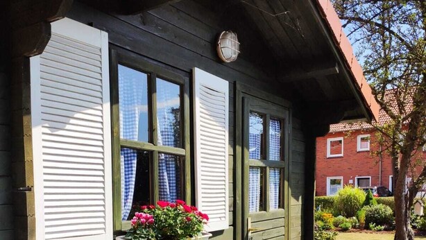 A wooden house with white shutters, a small porch, and a flower box with pink flowers. - Comfortable vacation home in a central location in Bad Zwischenahn with garden (Bad Zwischenahn)