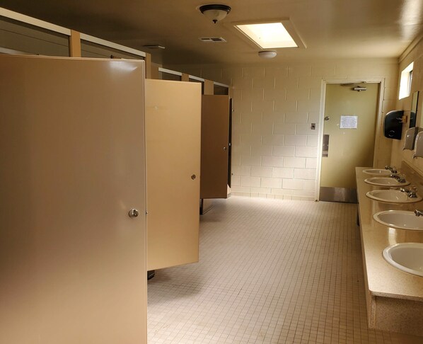 Baño público con múltiples cabinas, suelo de baldosas e lavamanos con dispensadores de jabón.