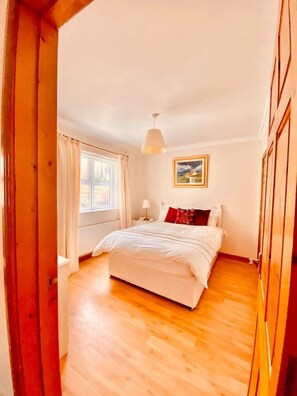 A bedroom with a wooden floor, a bed with white bedding and red pillows, a bedside table with a lamp, and a window with curtains. - Riverside Cottage, Ramelton, The Wild Atlantic Way (Ramelton)