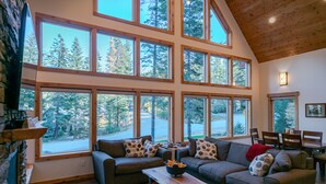 A spacious living room with a large window overlooking a forest, a cozy fireplace, and a comfortable seating area. - Mountain Retreat, steps from Skiing, Mountain Biking, and Hiking (Wenatchee)