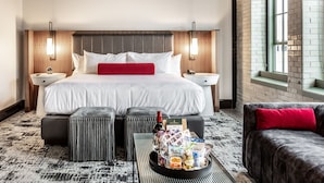 Et hotelværelse med en stor seng, to sengeborde, en rød pude, en bakke med snacks og en flaske, samt udsigt ud af vinduet.