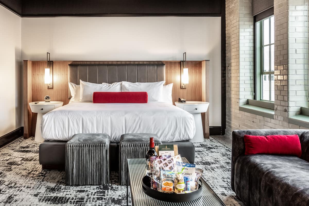 Ein Hotelzimmer mit einem großen Bett, zwei Nachttischen, einem roten Kissen, einer Ablage mit Snacks und einer Flasche sowie Blick nach draußen durch ein Fenster.