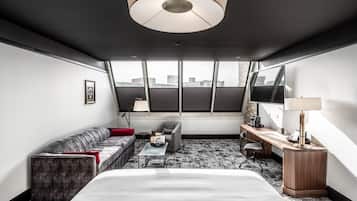 A modern hotel room with a large bed, a sofa, a desk, and a view of the city through large windows.