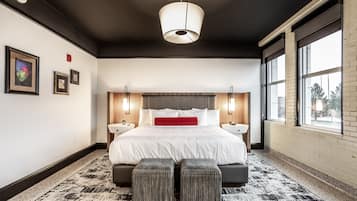 A modern hotel room with a large bed, two bedside tables, a red accent pillow, and a patterned rug.