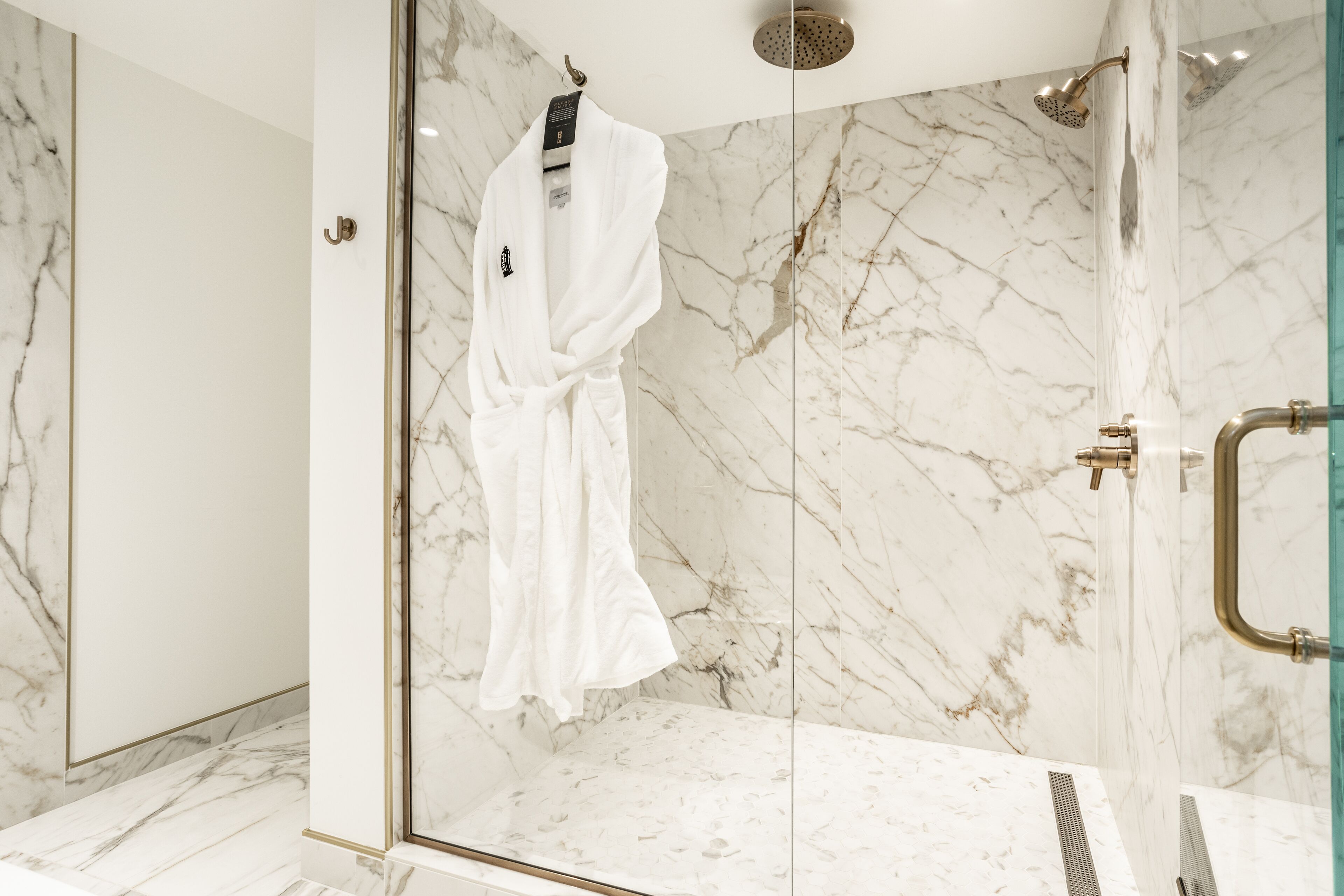 A modern bathroom with a glass-enclosed shower, marble walls, and a white robe hanging on a hook.