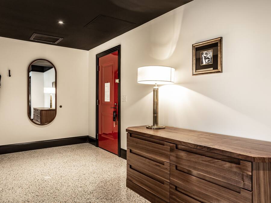 A hotel hallway with a wooden vanity, a mirror, a red door, and a framed picture on the wall.