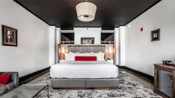 A modern hotel room with a large bed, a grey sofa, a patterned rug, and a ceiling light fixture.