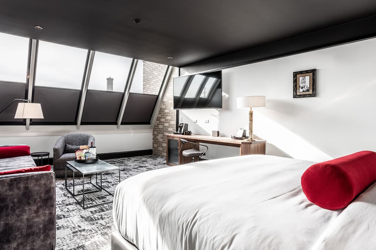 Ein modernes Hotelzimmer mit einem Bett, einem Schreibtisch, einem Sessel und einer Couch. Ein Fenster bietet Blick auf die Stadt.