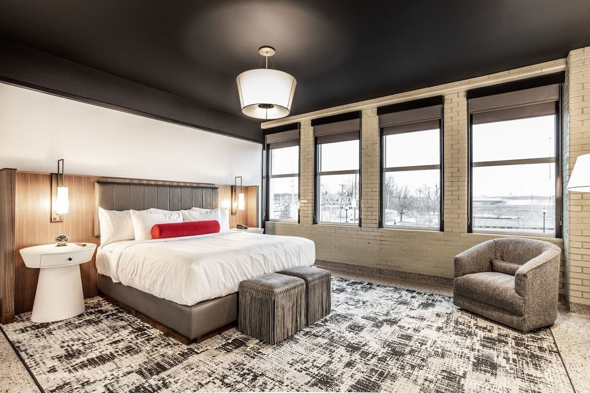 Ein modernes Hotelzimmer mit einem großen Bett, einem Nachttisch, einem Sessel und Blick durch große Fenster.