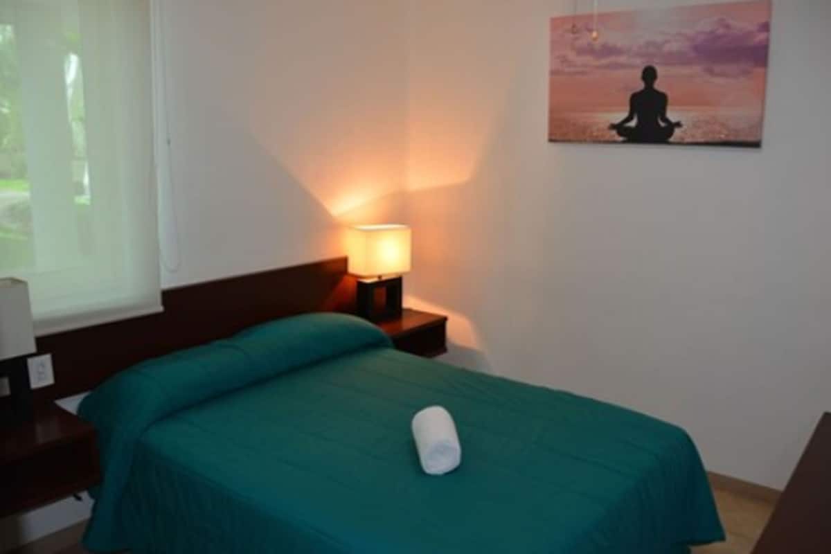 Ein Zimmer mit einem Bett, einem Nachttisch mit Lampe und einem Gemälde einer meditierenden Person.