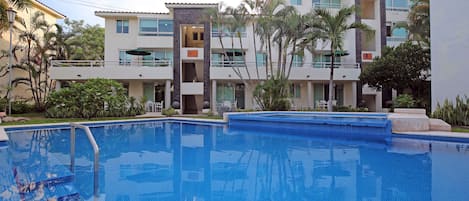 Una piscina con superficie de agua azul cristalina, rodeada por un edificio de varios pisos con balcones y un jardín ajardinado con palmeras.