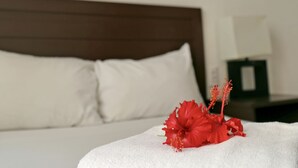 Habitación de hotel con una cama, almohadas blancas, una flor de hibisco rojo y un cuadro de un atardecer en la pared.