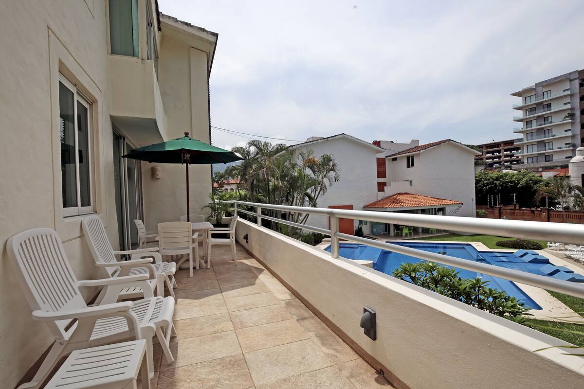 Ein Balkon mit weißen Stühlen, einem Tisch und einem grünen Sonnenschirm. Im Hintergrund ist ein Pool zu sehen.