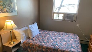 Une chambre à coucher avec un lit, une table de chevet avec une lampe, une fenêtre avec un climatiseur et un mur orné d’un tableau.