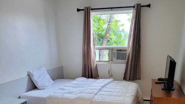 Un lit simple avec une couette blanche, une table de chevet en bois, une commode en bois et une fenêtre avec des rideaux.