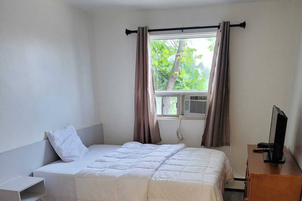 A single bed with a white duvet, a wooden nightstand, a wooden dresser, and a window with curtains.
