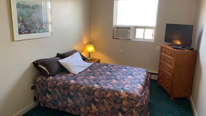 Une petite chambre à coucher, bien rangée, avec un lit, une commode, un téléviseur et une fenêtre équipée d’un appareil de climatisation.