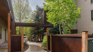 A wooden archway leading to a pathway surrounded by a wooden fence, with a tree and a building in the background.