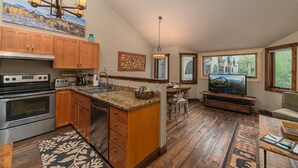 A spacious kitchen with wooden cabinets, a granite countertop, and a large island. There is a dining area with a wooden table and chairs, and a television mounted on the wall.