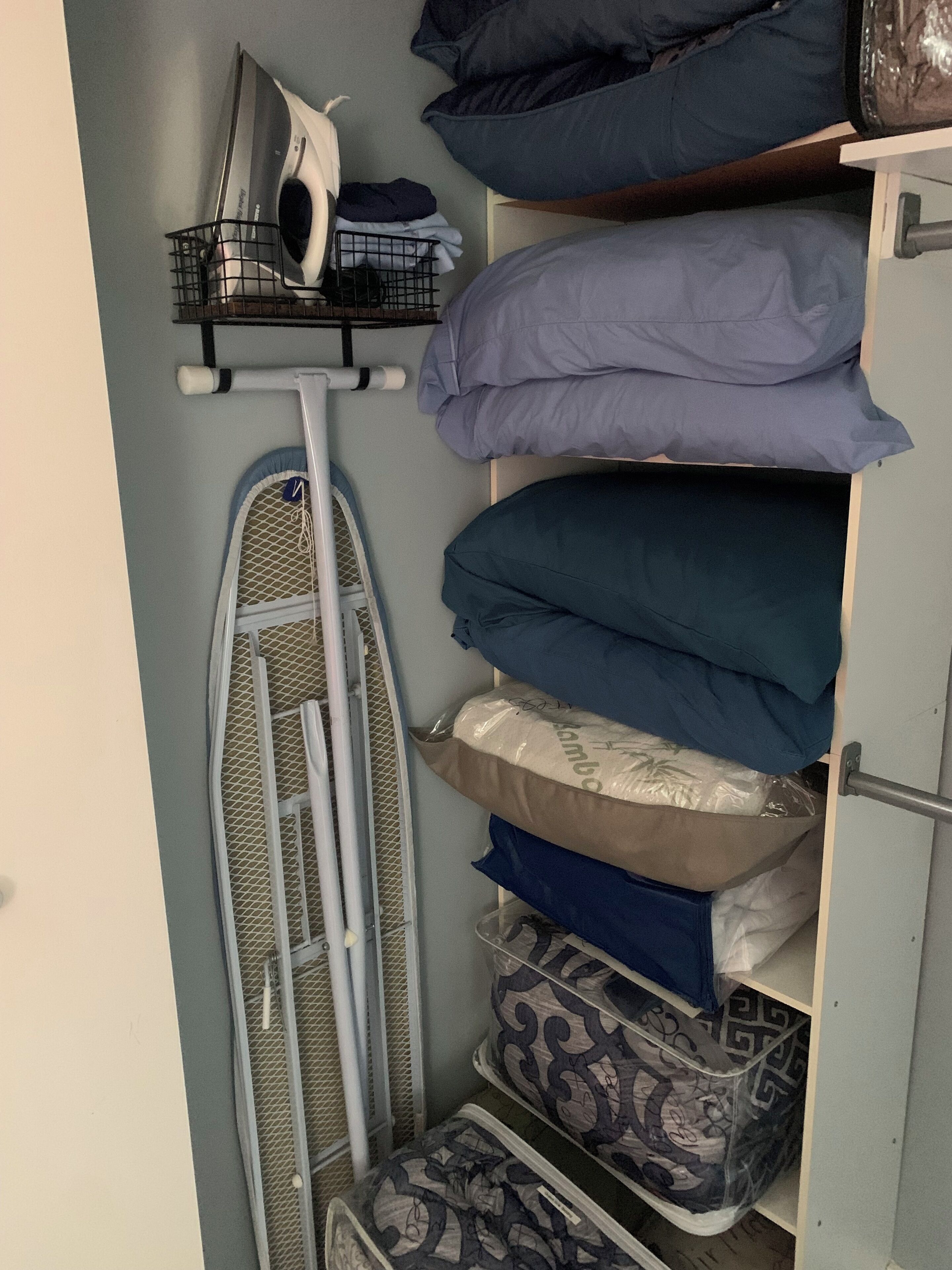 A storage area with shelves holding folded clothes, a hanging rack with an iron, and a mesh shelf with folded items.