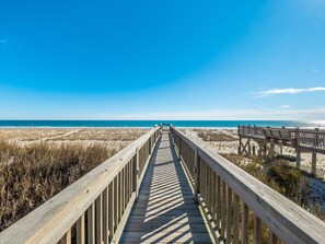 Eine Holzpromenade führt zu einem Strand mit Dünen und einem klaren, blauen Himmel.