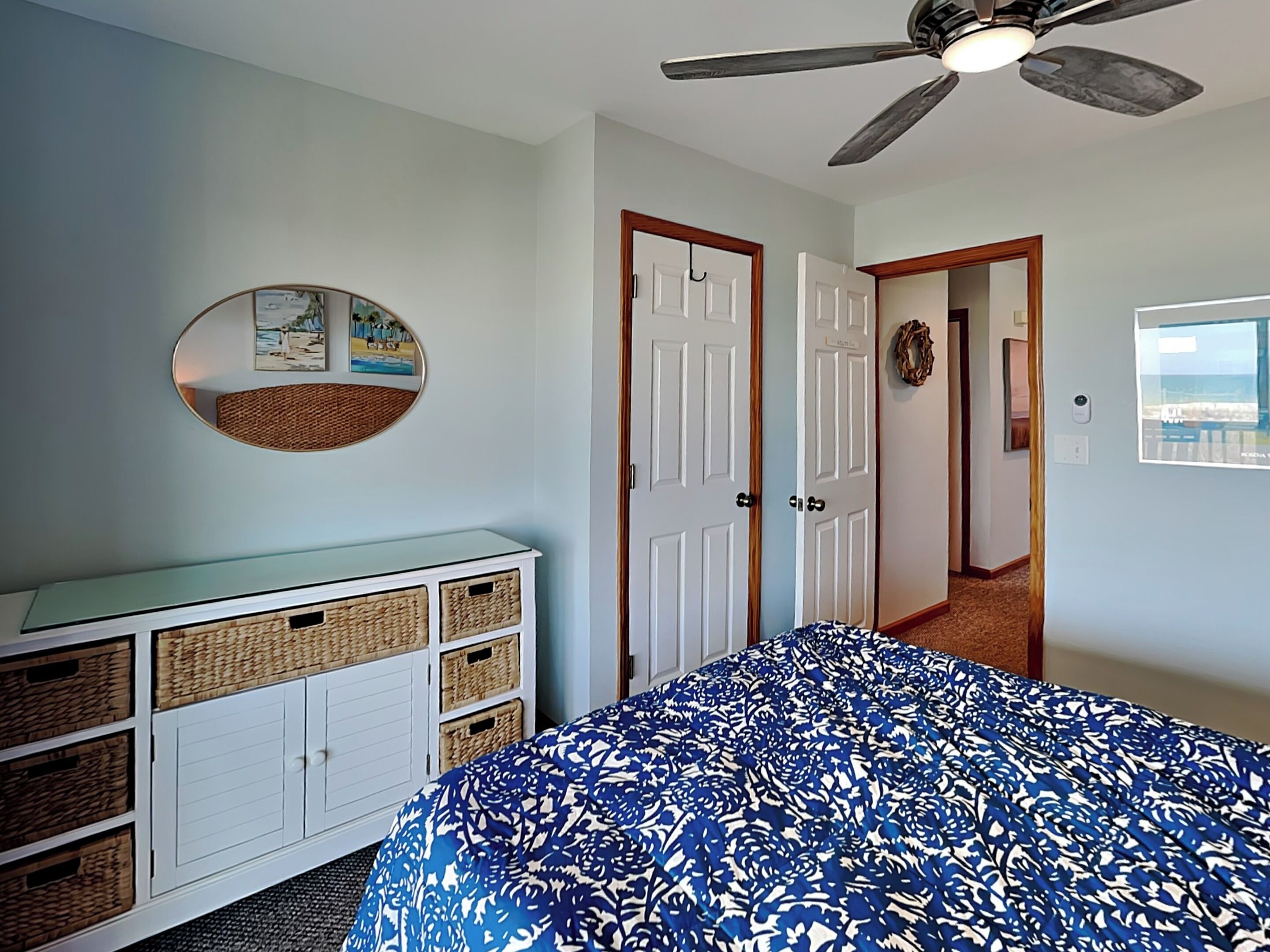 Un dormitorio con cama, ventilador de techo, cómoda con cestas, puerta decorada con un adorno y pared con un cuadro.