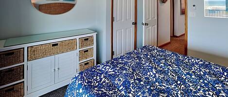 Un dormitorio con cama, ventilador de techo, cómoda con cestas, puerta decorada con un adorno y pared con un cuadro.
