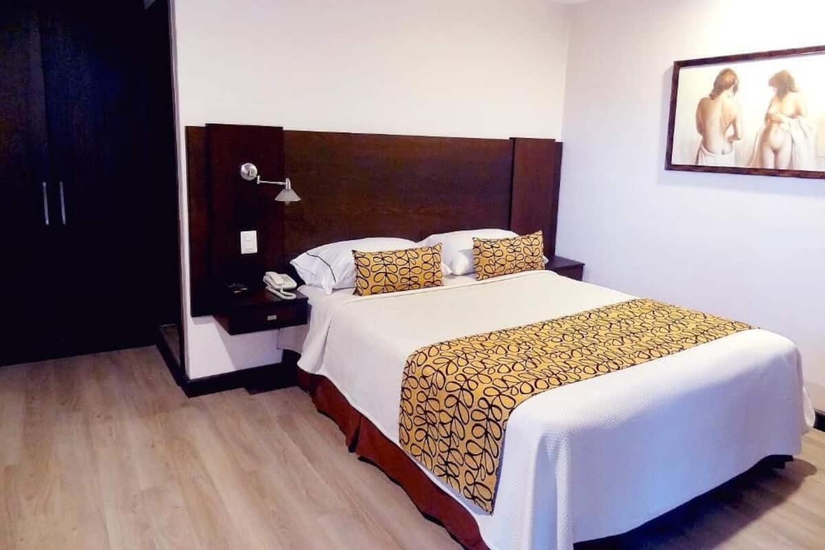 A hotel room with a large bed, a wooden headboard, a bedside table with a lamp, and a painting on the wall.