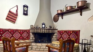 Ein gemütliches Wohnzimmer mit Kamin aus Stein, zwei Holzstühle mit bunten Kissen, einem roten Teppich mit Muster und einem Regal mit Kupferkochen.