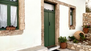 Ein traditionelles Gebäude mit einer grünen Tür, weißen Wänden und Steinverzierungen. Vor dem Eingang stehen Blumentöpfe und es gibt ein kleines Fenster mit Vorhang.