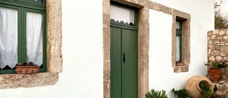 A traditional building with a green door, white walls, and stone accents. There are potted plants near the entrance and a small window with a curtain.