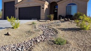 Une maison à deux étages avec un garage, une cour avant aménagée avec des rochers et de petites plantes, et un ciel bleu dégagé.