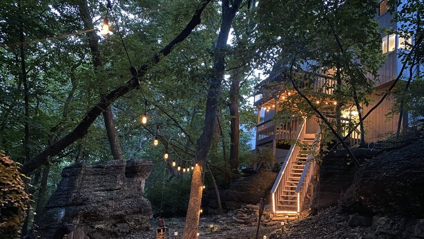 A rustic outdoor seating area with wooden chairs and a bench, surrounded by trees and a stone wall.