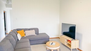 A modern living room with a grey sectional sofa, a small round ottoman, a TV on a wooden stand, and a white wall. - Dolphin Court - Park your car and walk to all that Robe has to offer (Robe)