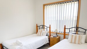A bedroom with two single beds, a wooden nightstand, and a window with blinds. - Dolphin Court - Park your car and walk to all that Robe has to offer (Robe)
