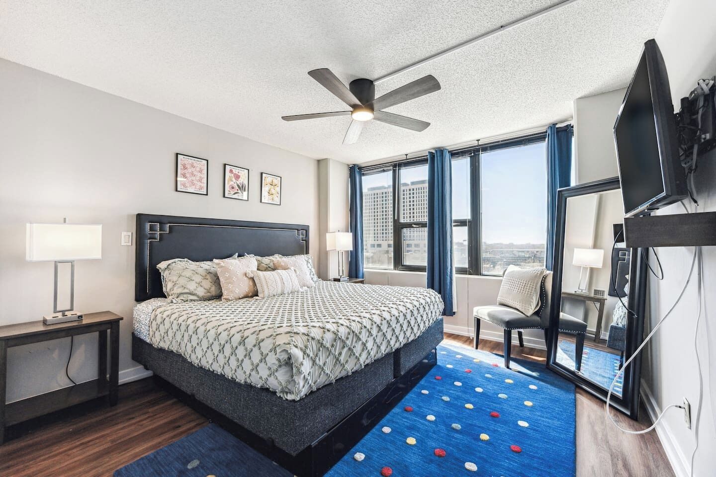 Una camera d'albergo moderna con un letto grande, una ventola a soffitto, una TV e vista sulla città attraverso ampie finestre.