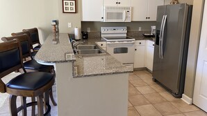 Una cocina con encimera de granito, armarios blancos, refrigerador de acero inoxidable, microondas y estufa.