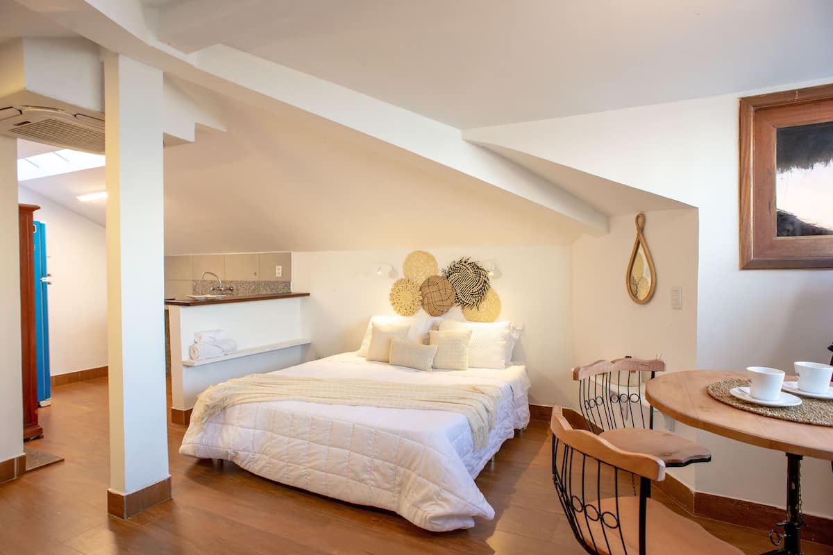 A bedroom with a large bed, a wooden chair, a small table with cups, and a wall clock.