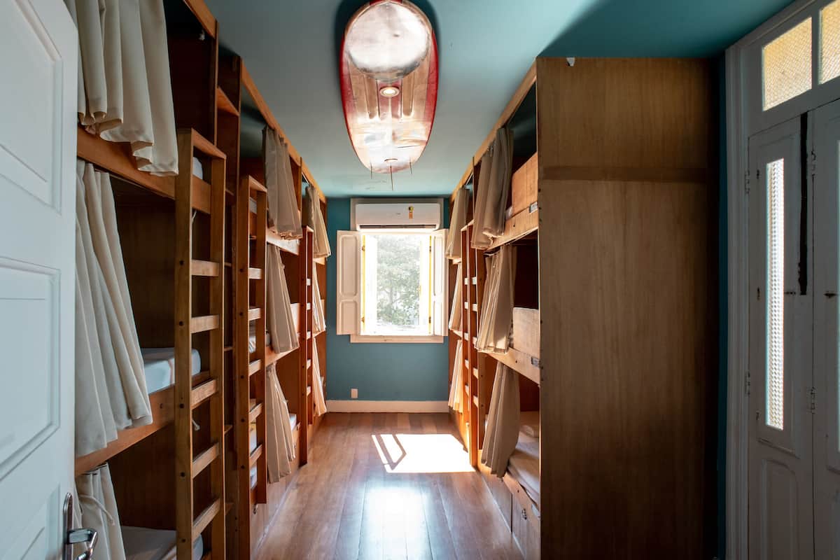 A narrow hallway with wooden shelving units on both sides, a window at the end, and a surfboard-shaped light fixture on the ceiling.