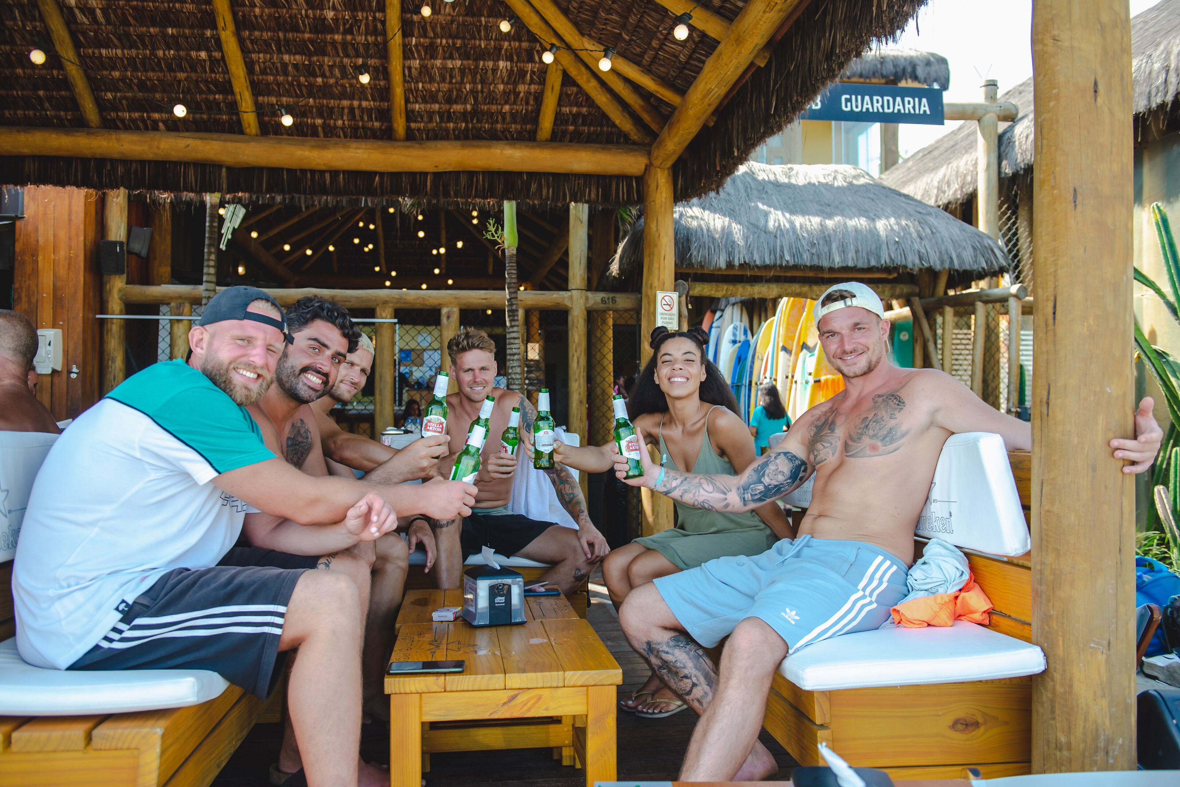 Een groep mensen zit aan een houten tafel onder een rieten dak, met bierflesjes in hun handen.